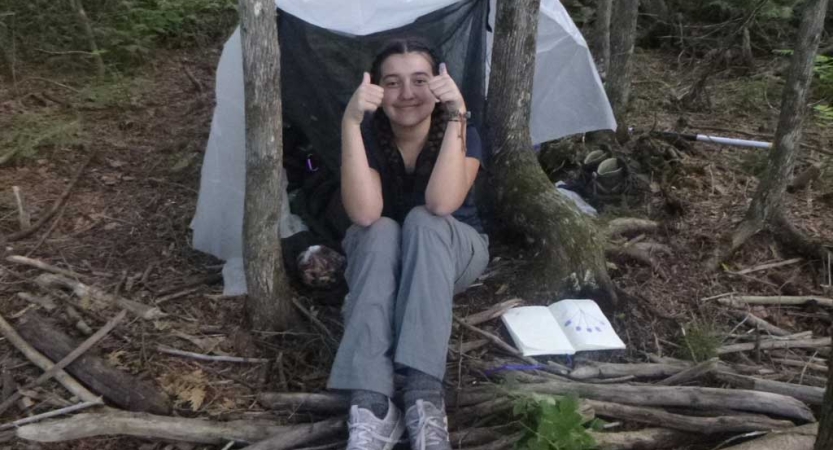 A person sits beside a tarp shelter in a wooded area and gives two thumbs up